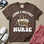 A Labor and Delivery nurse graphic featuring a large beige tote bag with a leopard print pocket, a stethoscope draped over it, and a large iced coffee cup with a matching leopard print sleeve, all surrounded by white text that reads LABOR & DELIVERY curved above and NURSE below.