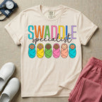 A cute neonatal and postpartum nurse graphic featuring the word SWADDLE in bright multicolored block letters above the word specialist in a white cursive font, positioned over a row of six diverse babies wrapped in brightly colored swaddles.