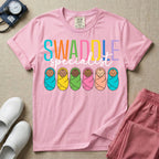 A cute neonatal and postpartum nurse graphic featuring the word SWADDLE in bright multicolored block letters above the word specialist in a white cursive font, positioned over a row of six diverse babies wrapped in brightly colored swaddles.