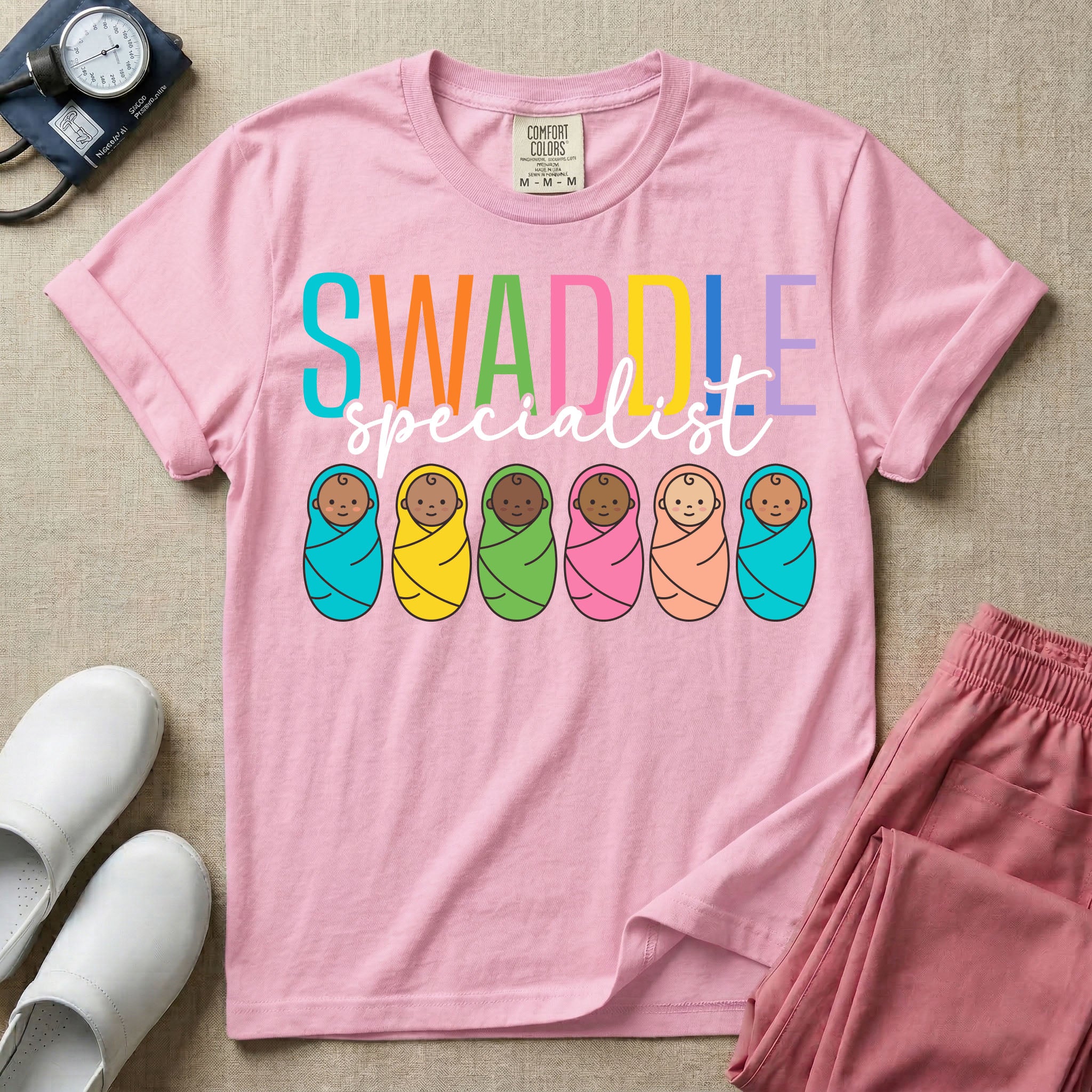 A cute neonatal and postpartum nurse graphic featuring the word SWADDLE in bright multicolored block letters above the word specialist in a white cursive font, positioned over a row of six diverse babies wrapped in brightly colored swaddles.