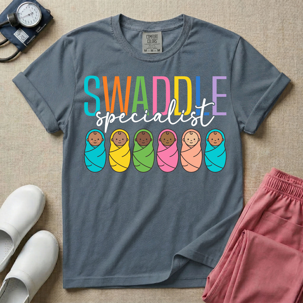 A cute neonatal and postpartum nurse graphic featuring the word SWADDLE in bright multicolored block letters above the word specialist in a white cursive font, positioned over a row of six diverse babies wrapped in brightly colored swaddles.