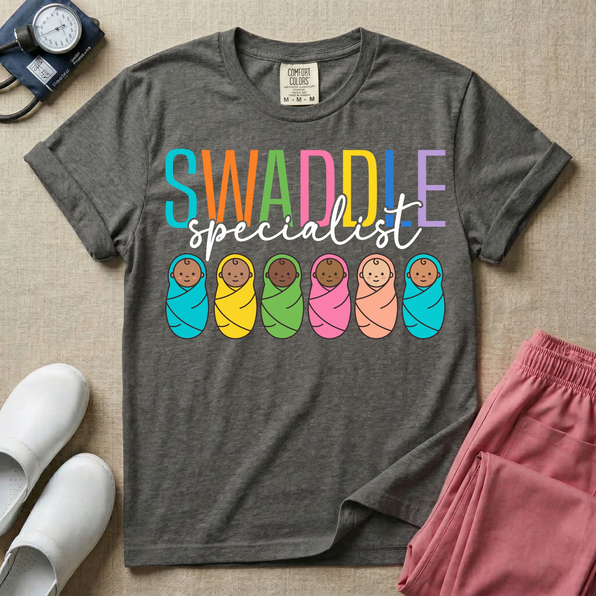 A cute neonatal and postpartum nurse graphic featuring the word SWADDLE in bright multicolored block letters above the word specialist in a white cursive font, positioned over a row of six diverse babies wrapped in brightly colored swaddles.
