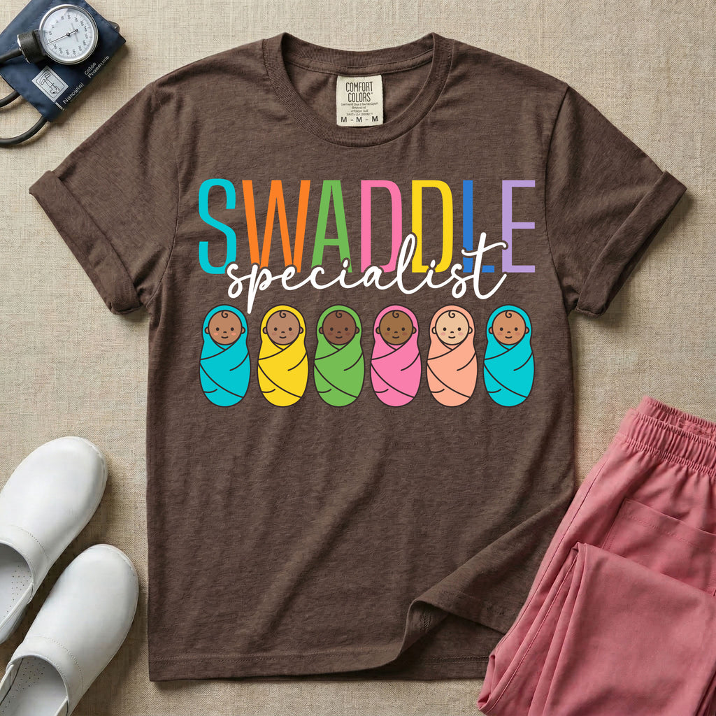 A cute neonatal and postpartum nurse graphic featuring the word SWADDLE in bright multicolored block letters above the word specialist in a white cursive font, positioned over a row of six diverse babies wrapped in brightly colored swaddles.