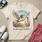 A humorous Labor and Delivery graphic featuring a storybook illustration of a frog dressed as a knight wielding a sword, riding on the back of a flying stork that is carrying a sleeping baby in a bundle, soaring over a fantasy landscape with a castle at sunrise, accompanied by text below that reads We deliver at dawn.