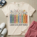 A Labor and Delivery nurse graphic featuring a row of standing books with colorful wildflowers blooming from the top pages. The book spines feature humorous titles reading CATCHIN' BABIES, PITOCIN 101, CERVIX CHECKS, CROWNING GLORY, POSTPARTUM PRO, PUSH, BREATHE, REPEAT, FETAL HEART TONES, IN THE LABORHOOD, and ALMOST 10CM (AGAIN), with the text LABOR & DELIVERY NURSE written directly below.