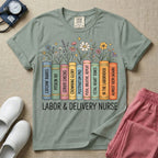 A Labor and Delivery nurse graphic featuring a row of standing books with colorful wildflowers blooming from the top pages. The book spines feature humorous titles reading CATCHIN' BABIES, PITOCIN 101, CERVIX CHECKS, CROWNING GLORY, POSTPARTUM PRO, PUSH, BREATHE, REPEAT, FETAL HEART TONES, IN THE LABORHOOD, and ALMOST 10CM (AGAIN), with the text LABOR & DELIVERY NURSE written directly below.
