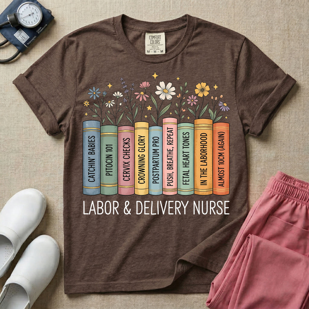 A Labor and Delivery nurse graphic featuring a row of standing books with colorful wildflowers blooming from the top pages. The book spines feature humorous titles reading CATCHIN' BABIES, PITOCIN 101, CERVIX CHECKS, CROWNING GLORY, POSTPARTUM PRO, PUSH, BREATHE, REPEAT, FETAL HEART TONES, IN THE LABORHOOD, and ALMOST 10CM (AGAIN), with the text LABOR & DELIVERY NURSE written directly below.