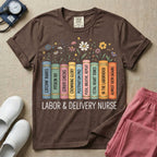 A Labor and Delivery nurse graphic featuring a row of standing books with colorful wildflowers blooming from the top pages. The book spines feature humorous titles reading CATCHIN' BABIES, PITOCIN 101, CERVIX CHECKS, CROWNING GLORY, POSTPARTUM PRO, PUSH, BREATHE, REPEAT, FETAL HEART TONES, IN THE LABORHOOD, and ALMOST 10CM (AGAIN), with the text LABOR & DELIVERY NURSE written directly below.