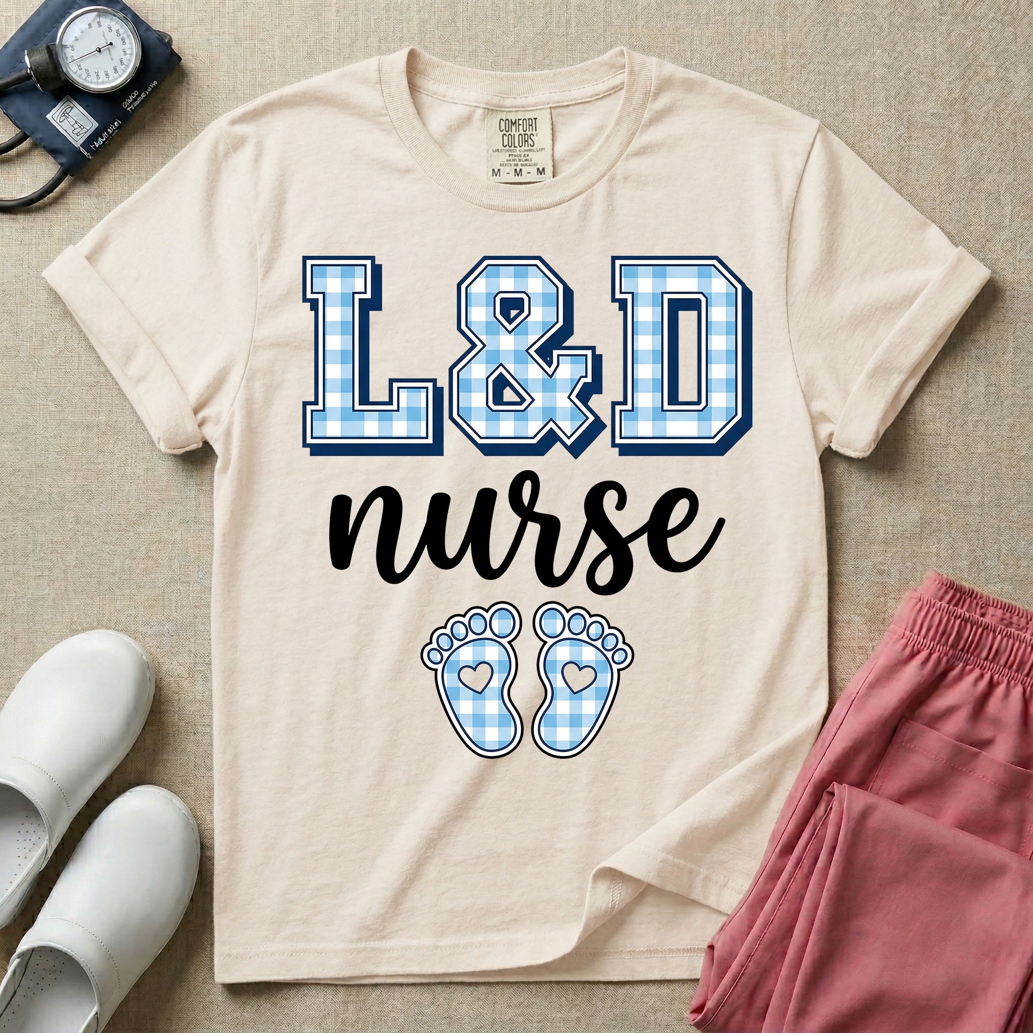 A graphic t-shirt for a Labor and Delivery unit featuring the text 'L&D nurse' with a light blue gingham pattern inside the block letters, along with matching blue gingham baby footprints featuring small heart cutouts.