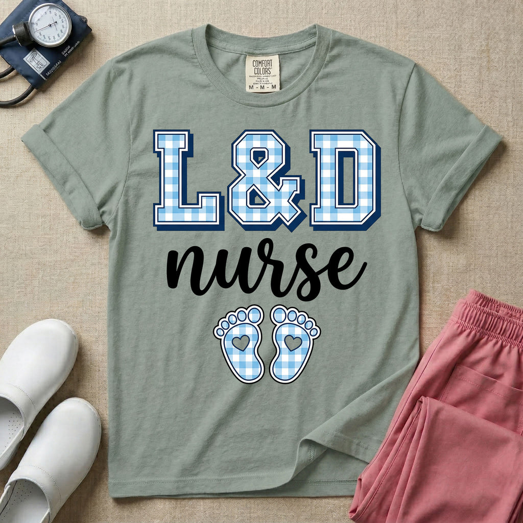 A graphic t-shirt for a Labor and Delivery unit featuring the text 'L&D nurse' with a light blue gingham pattern inside the block letters, along with matching blue gingham baby footprints featuring small heart cutouts.