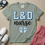 A graphic t-shirt for a Labor and Delivery unit featuring the text 'L&D nurse' with a light blue gingham pattern inside the block letters, along with matching blue gingham baby footprints featuring small heart cutouts.