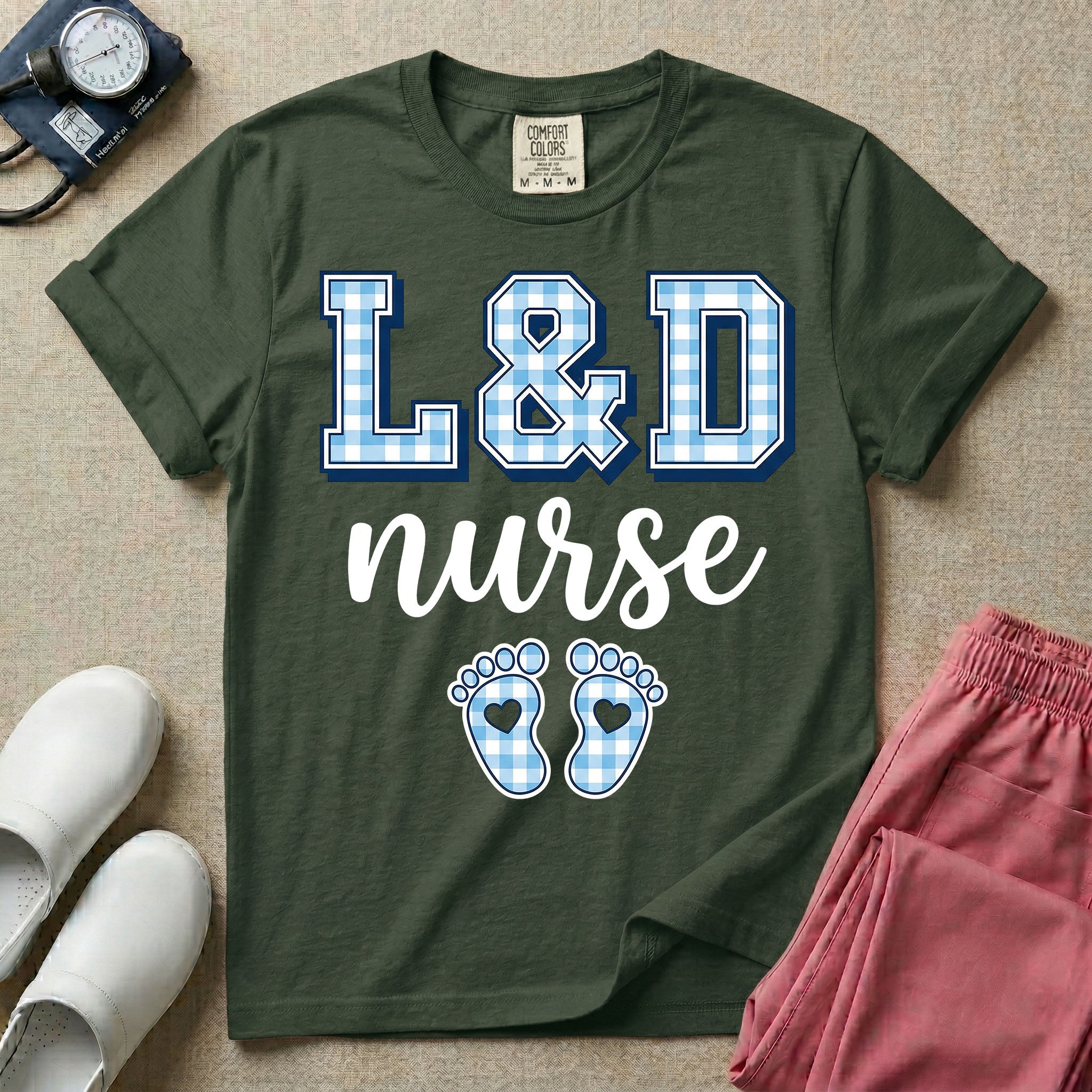 A graphic t-shirt for a Labor and Delivery unit featuring the text 'L&D nurse' with a light blue gingham pattern inside the block letters, along with matching blue gingham baby footprints featuring small heart cutouts.
