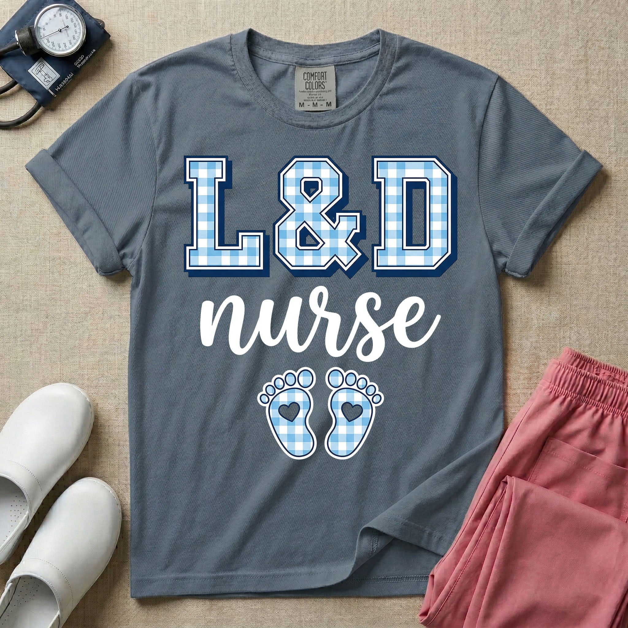 A graphic t-shirt for a Labor and Delivery unit featuring the text 'L&D nurse' with a light blue gingham pattern inside the block letters, along with matching blue gingham baby footprints featuring small heart cutouts.