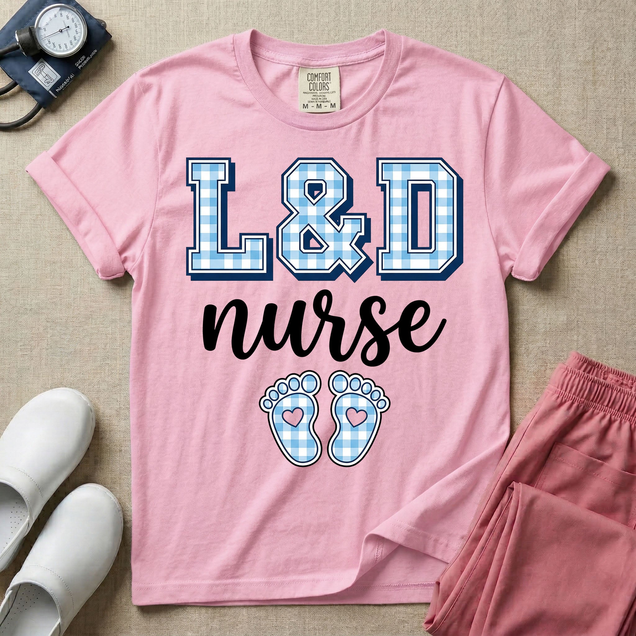 A graphic t-shirt for a Labor and Delivery unit featuring the text 'L&D nurse' with a light blue gingham pattern inside the block letters, along with matching blue gingham baby footprints featuring small heart cutouts.