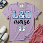 A graphic t-shirt for a Labor and Delivery unit featuring the text 'L&D nurse' with a light blue gingham pattern inside the block letters, along with matching blue gingham baby footprints featuring small heart cutouts.