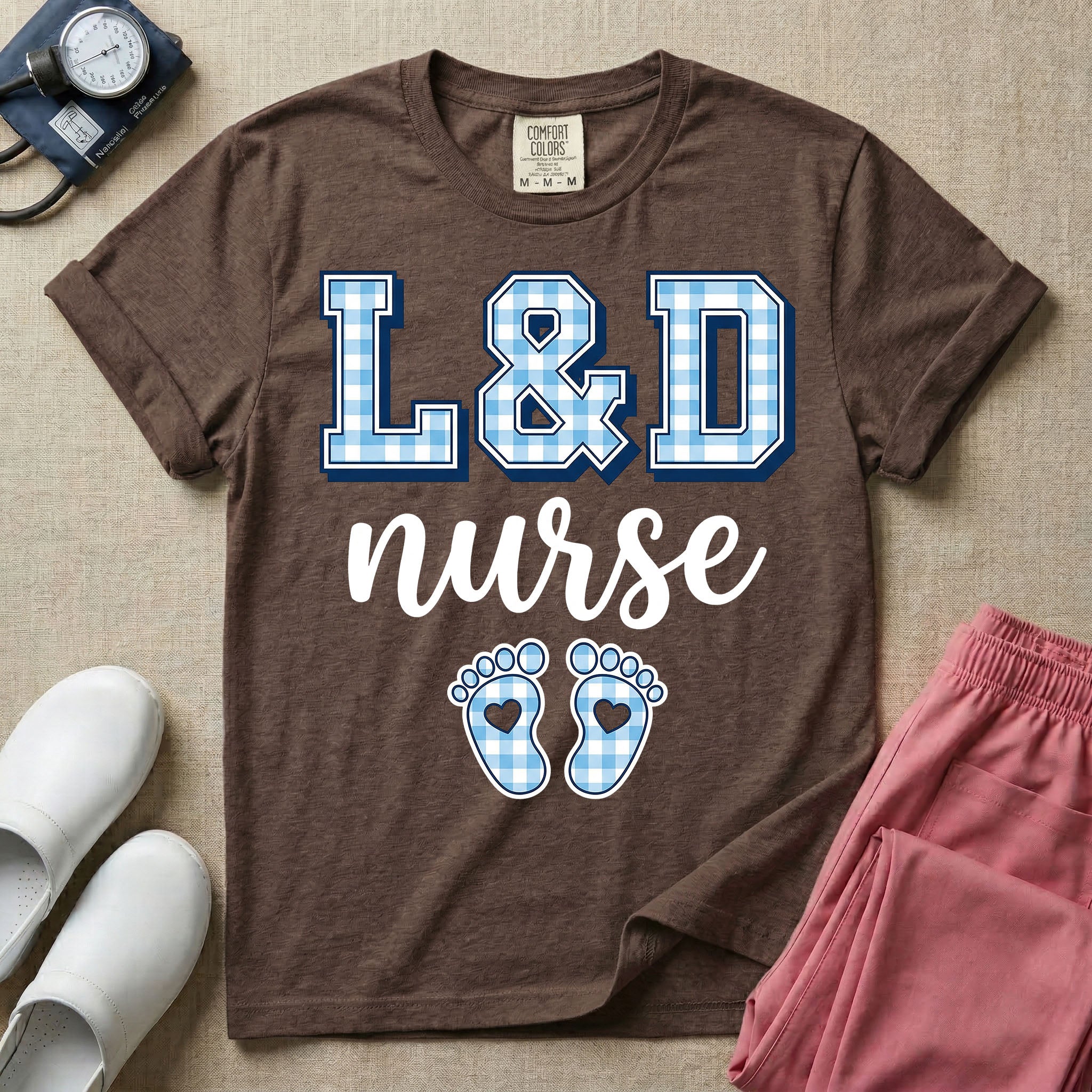 A graphic t-shirt for a Labor and Delivery unit featuring the text 'L&D nurse' with a light blue gingham pattern inside the block letters, along with matching blue gingham baby footprints featuring small heart cutouts.