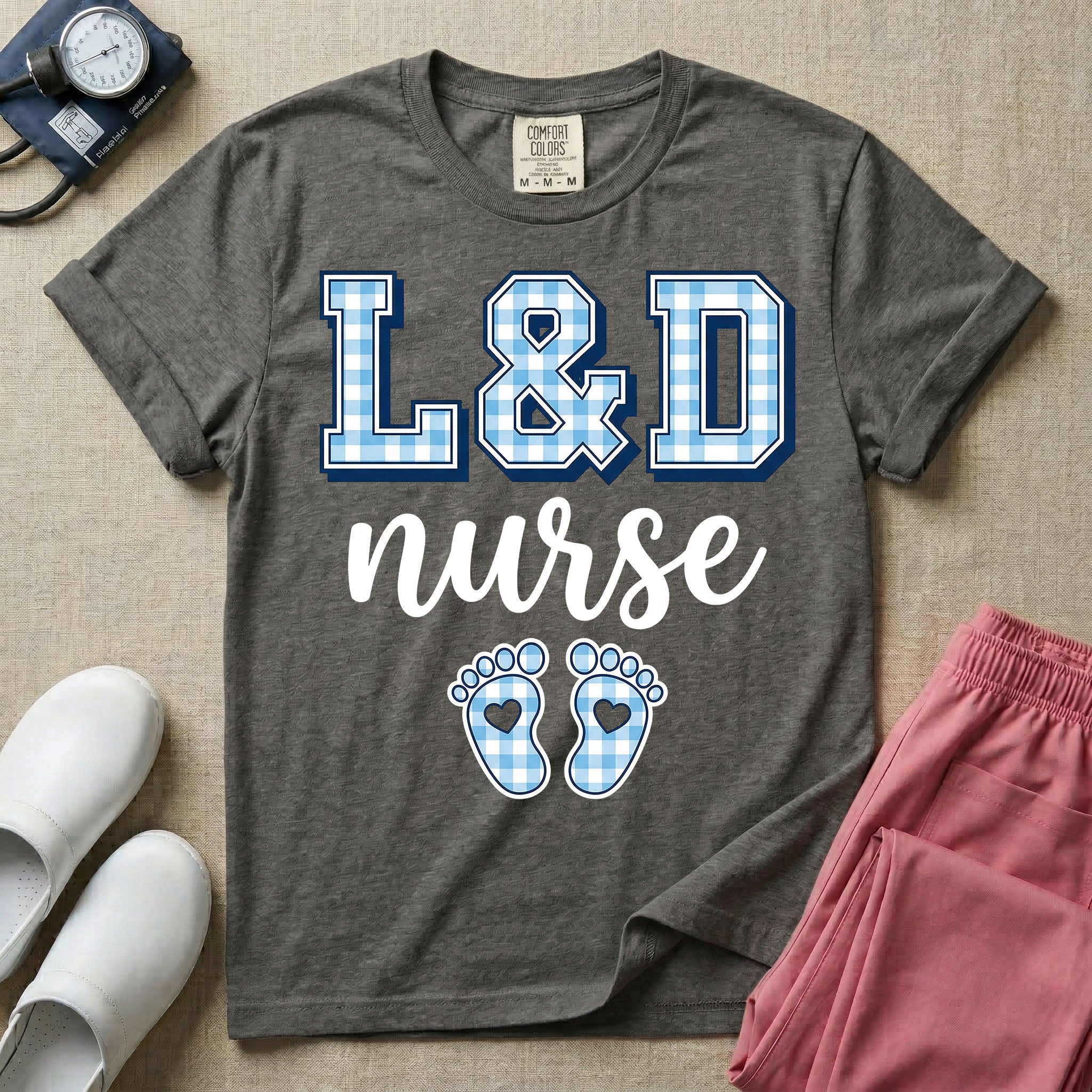 A graphic t-shirt for a Labor and Delivery unit featuring the text 'L&D nurse' with a light blue gingham pattern inside the block letters, along with matching blue gingham baby footprints featuring small heart cutouts.