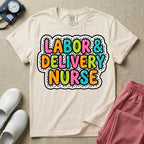 A graphic t-shirt for a Labor and Delivery unit featuring the text 'LABOR & DELIVERY NURSE' in bright, multicolored bubble lettering with white dots, framed by a contrasting black and white spotted background pattern.