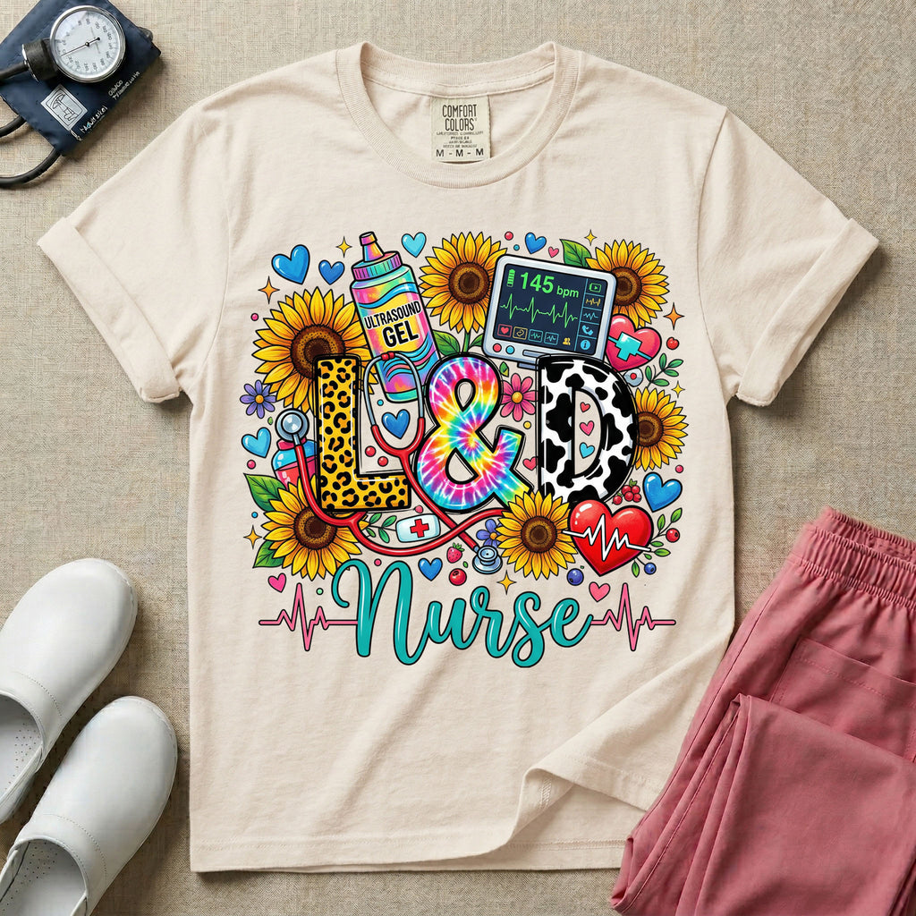 A colorful graphic t-shirt for a Labor and Delivery unit featuring the text "L&D" in leopard print, tie-dye, and cow print letters above the word "Nurse" in teal script, surrounded by detailed illustrations of sunflowers, a rainbow ultrasound gel bottle, a fetal monitor reading 145 bpm, a red stethoscope, and various hearts.