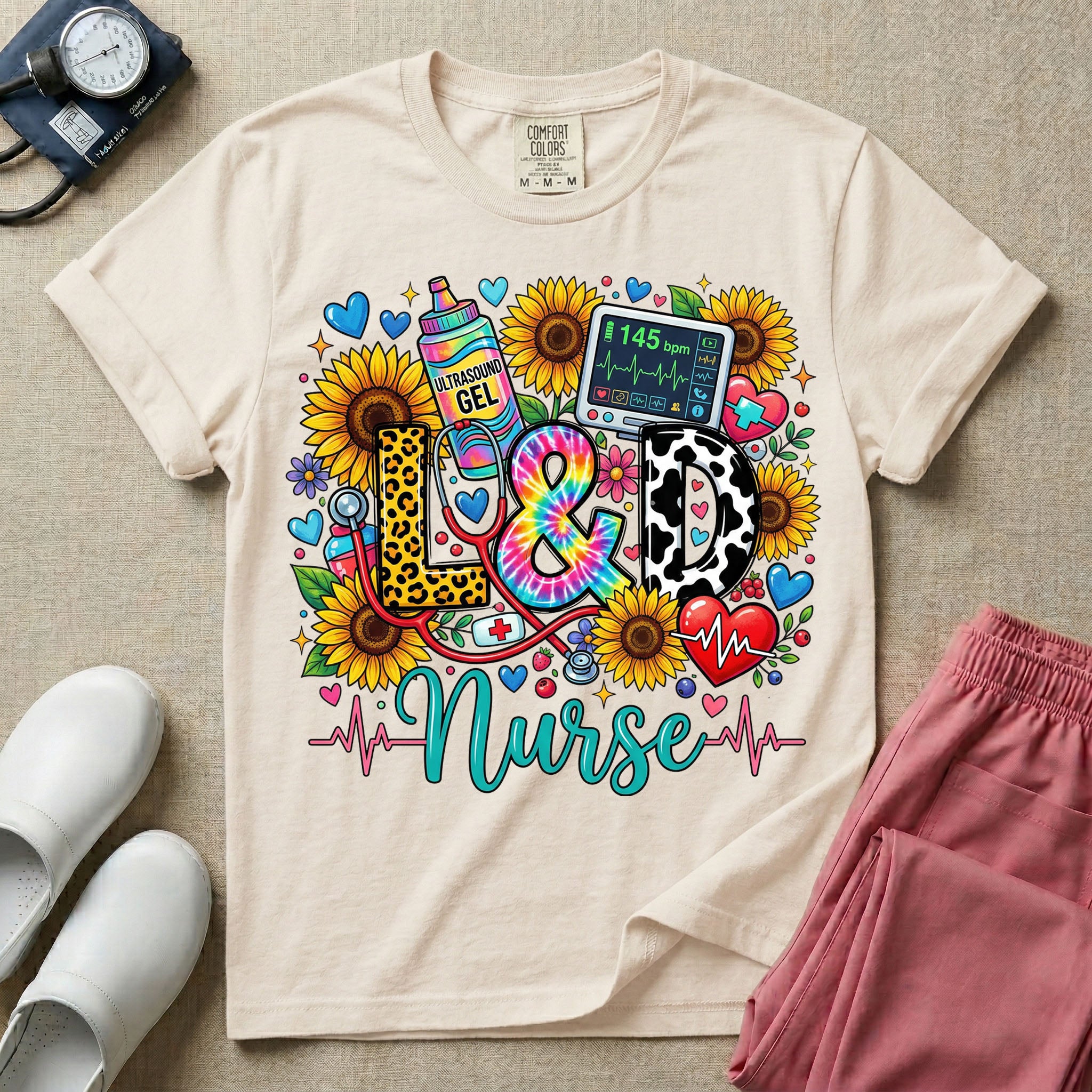A colorful graphic t-shirt for a Labor and Delivery unit featuring the text "L&D" in leopard print, tie-dye, and cow print letters above the word "Nurse" in teal script, surrounded by detailed illustrations of sunflowers, a rainbow ultrasound gel bottle, a fetal monitor reading 145 bpm, a red stethoscope, and various hearts.