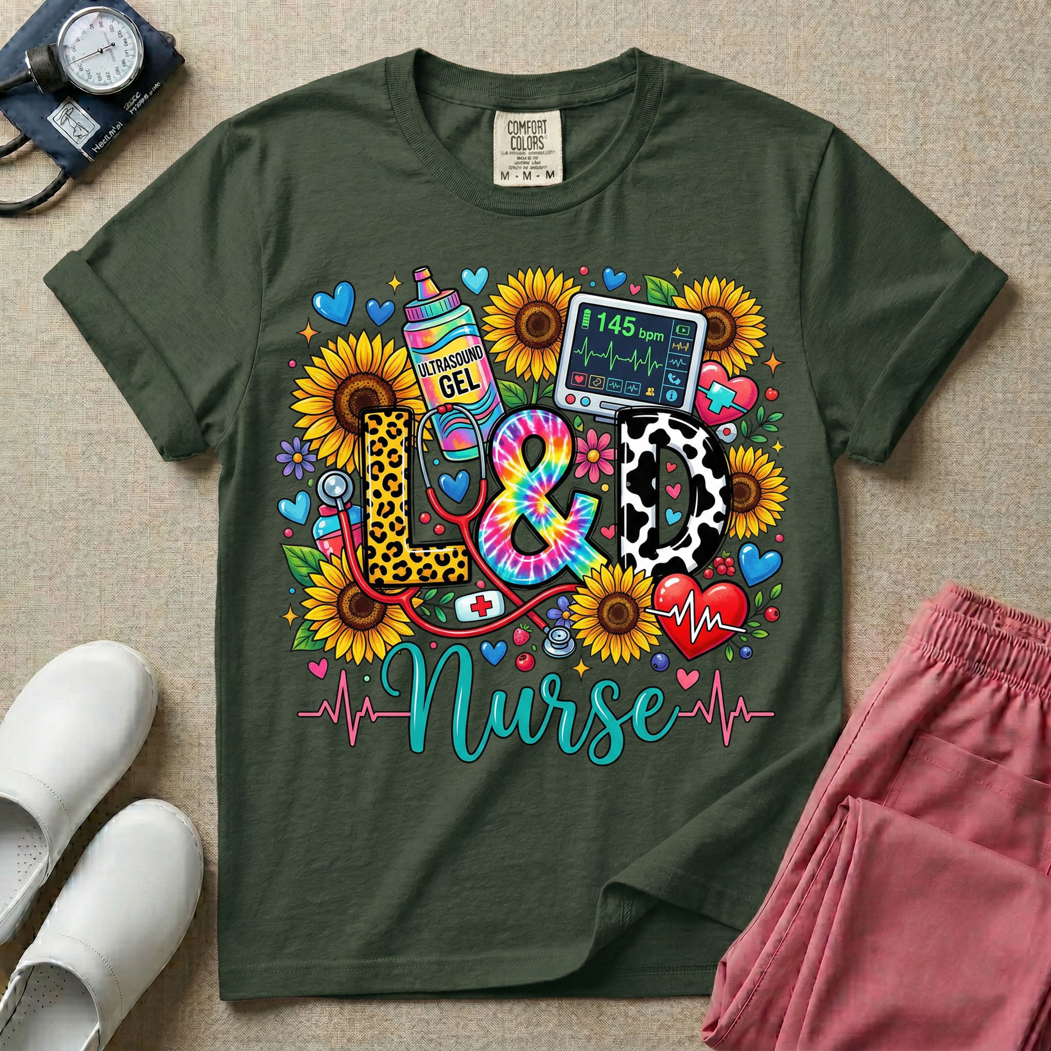 A colorful graphic t-shirt for a Labor and Delivery unit featuring the text "L&D" in leopard print, tie-dye, and cow print letters above the word "Nurse" in teal script, surrounded by detailed illustrations of sunflowers, a rainbow ultrasound gel bottle, a fetal monitor reading 145 bpm, a red stethoscope, and various hearts.