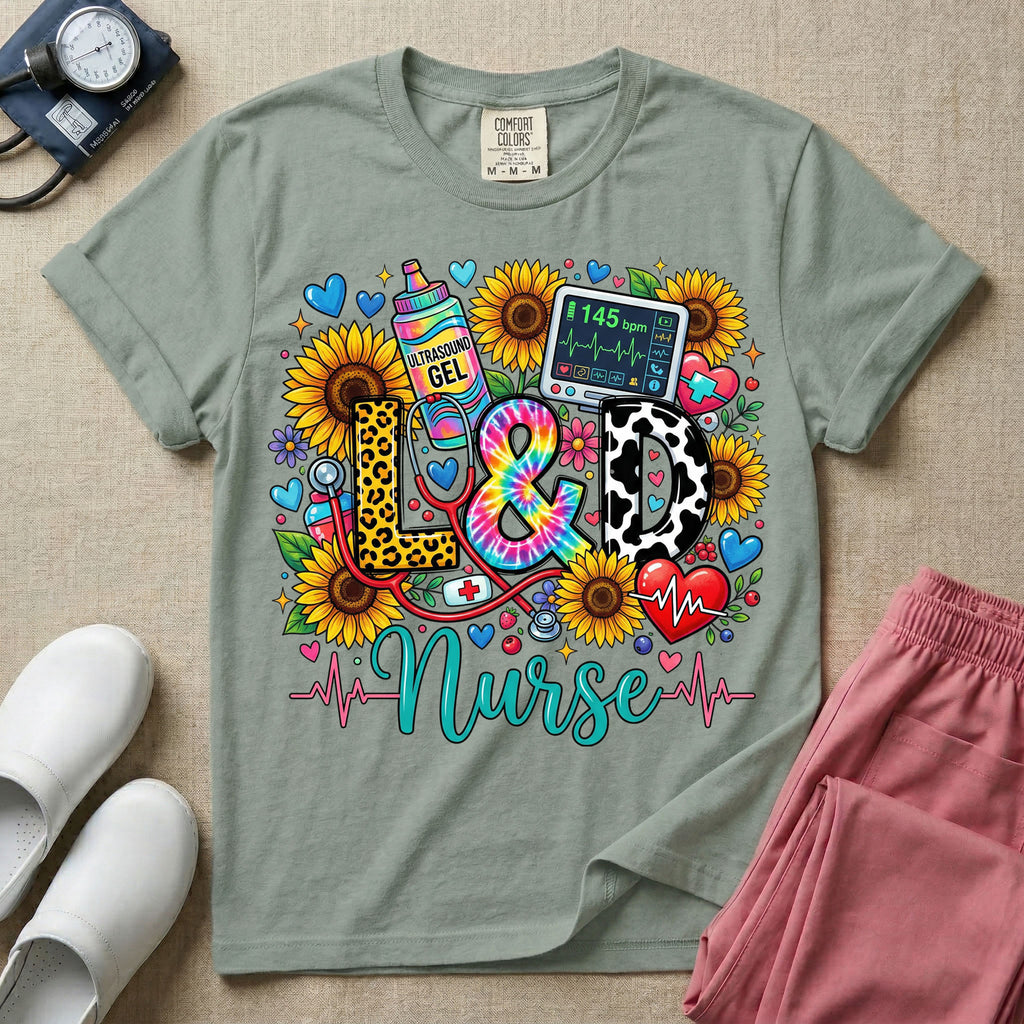A colorful graphic t-shirt for a Labor and Delivery unit featuring the text "L&D" in leopard print, tie-dye, and cow print letters above the word "Nurse" in teal script, surrounded by detailed illustrations of sunflowers, a rainbow ultrasound gel bottle, a fetal monitor reading 145 bpm, a red stethoscope, and various hearts.