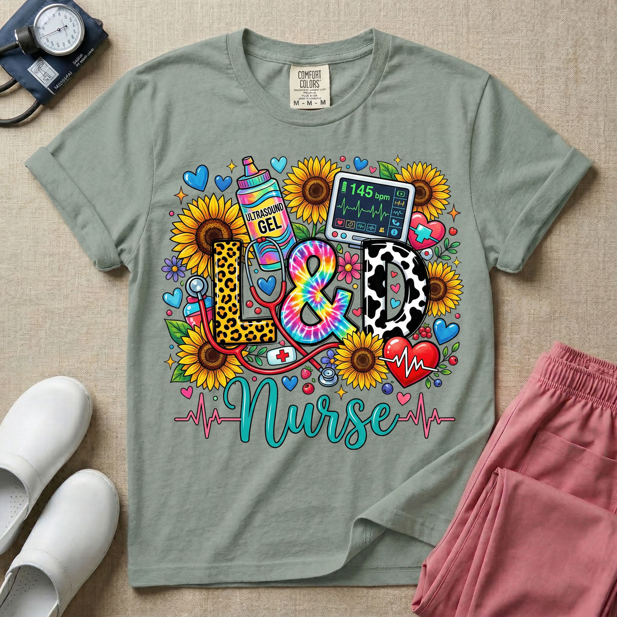A colorful graphic t-shirt for a Labor and Delivery unit featuring the text "L&D" in leopard print, tie-dye, and cow print letters above the word "Nurse" in teal script, surrounded by detailed illustrations of sunflowers, a rainbow ultrasound gel bottle, a fetal monitor reading 145 bpm, a red stethoscope, and various hearts.