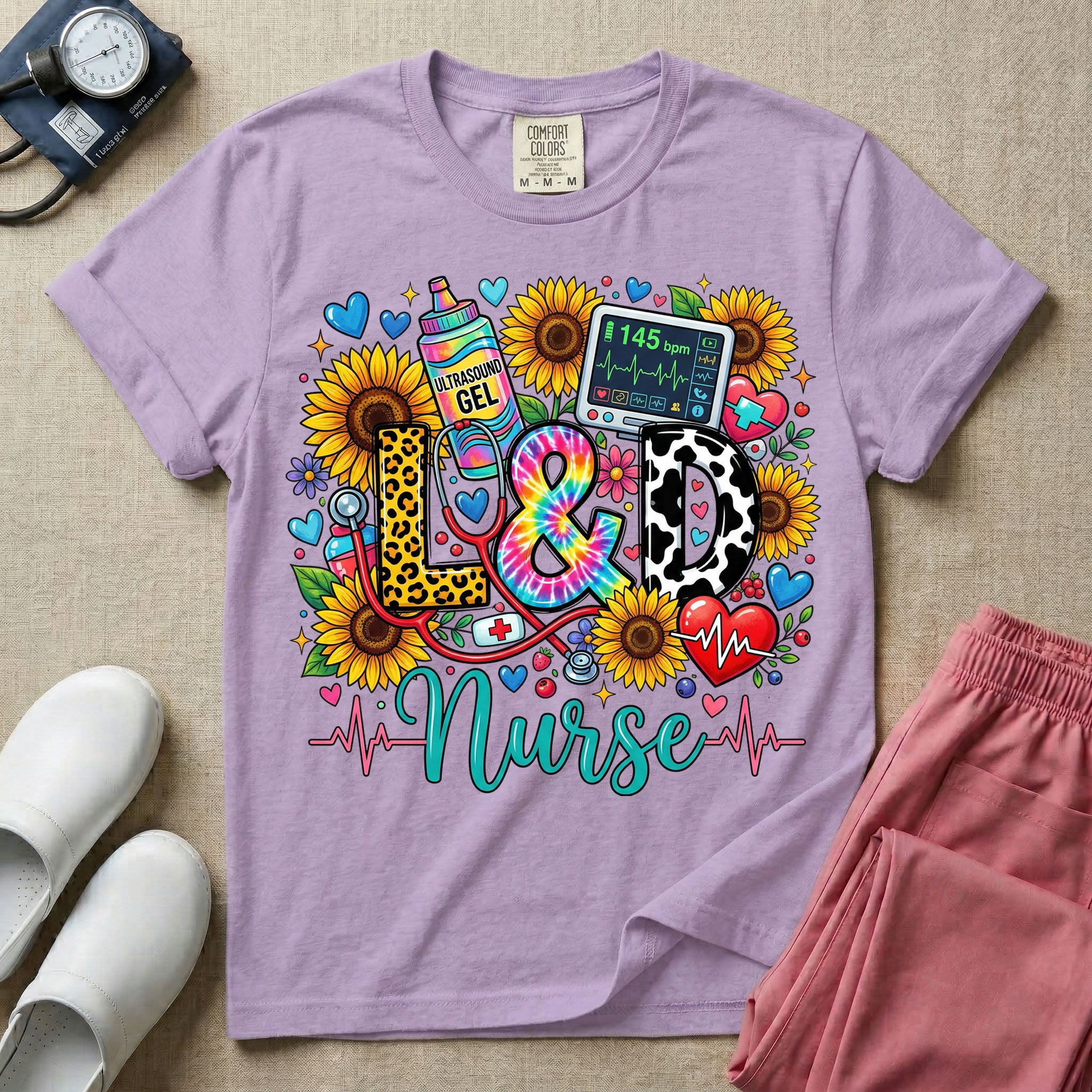 A colorful graphic t-shirt for a Labor and Delivery unit featuring the text "L&D" in leopard print, tie-dye, and cow print letters above the word "Nurse" in teal script, surrounded by detailed illustrations of sunflowers, a rainbow ultrasound gel bottle, a fetal monitor reading 145 bpm, a red stethoscope, and various hearts.