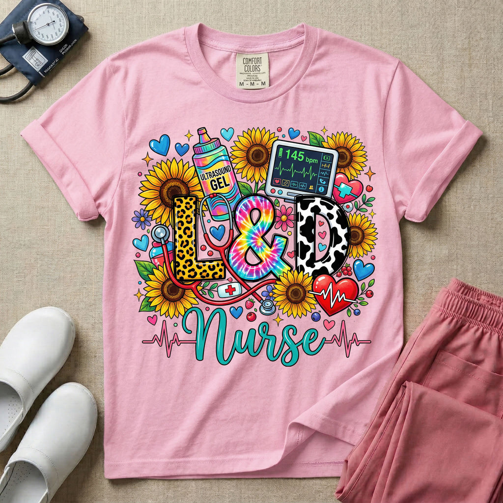 A colorful graphic t-shirt for a Labor and Delivery unit featuring the text "L&D" in leopard print, tie-dye, and cow print letters above the word "Nurse" in teal script, surrounded by detailed illustrations of sunflowers, a rainbow ultrasound gel bottle, a fetal monitor reading 145 bpm, a red stethoscope, and various hearts.