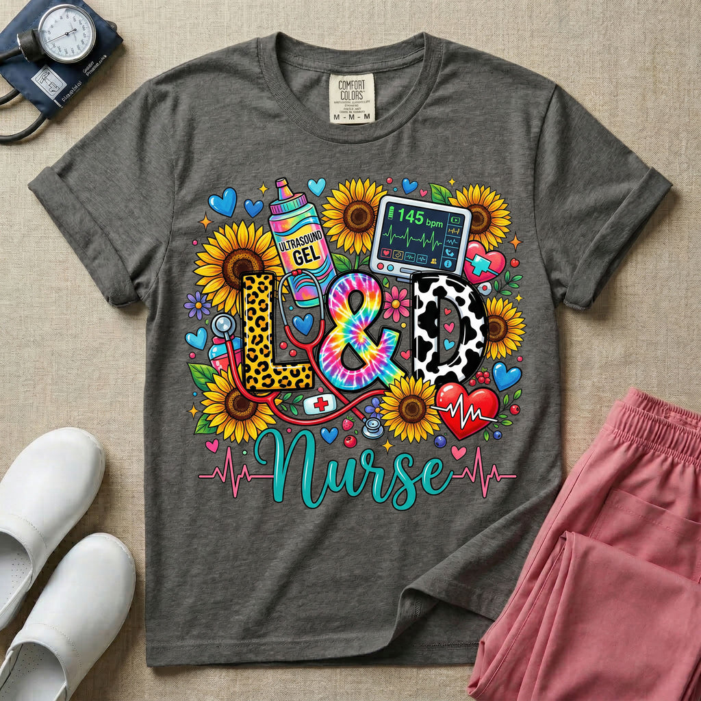 A colorful graphic t-shirt for a Labor and Delivery unit featuring the text "L&D" in leopard print, tie-dye, and cow print letters above the word "Nurse" in teal script, surrounded by detailed illustrations of sunflowers, a rainbow ultrasound gel bottle, a fetal monitor reading 145 bpm, a red stethoscope, and various hearts.