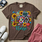 A colorful graphic t-shirt for a Labor and Delivery unit featuring the text "L&D" in leopard print, tie-dye, and cow print letters above the word "Nurse" in teal script, surrounded by detailed illustrations of sunflowers, a rainbow ultrasound gel bottle, a fetal monitor reading 145 bpm, a red stethoscope, and various hearts.