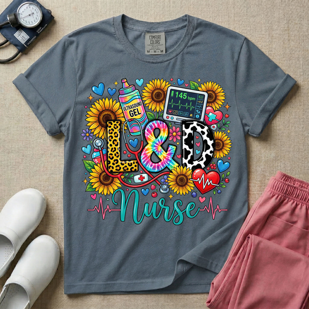 A colorful graphic t-shirt for a Labor and Delivery unit featuring the text "L&D" in leopard print, tie-dye, and cow print letters above the word "Nurse" in teal script, surrounded by detailed illustrations of sunflowers, a rainbow ultrasound gel bottle, a fetal monitor reading 145 bpm, a red stethoscope, and various hearts.