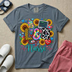 A colorful graphic t-shirt for a Labor and Delivery unit featuring the text "L&D" in leopard print, tie-dye, and cow print letters above the word "Nurse" in teal script, surrounded by detailed illustrations of sunflowers, a rainbow ultrasound gel bottle, a fetal monitor reading 145 bpm, a red stethoscope, and various hearts.