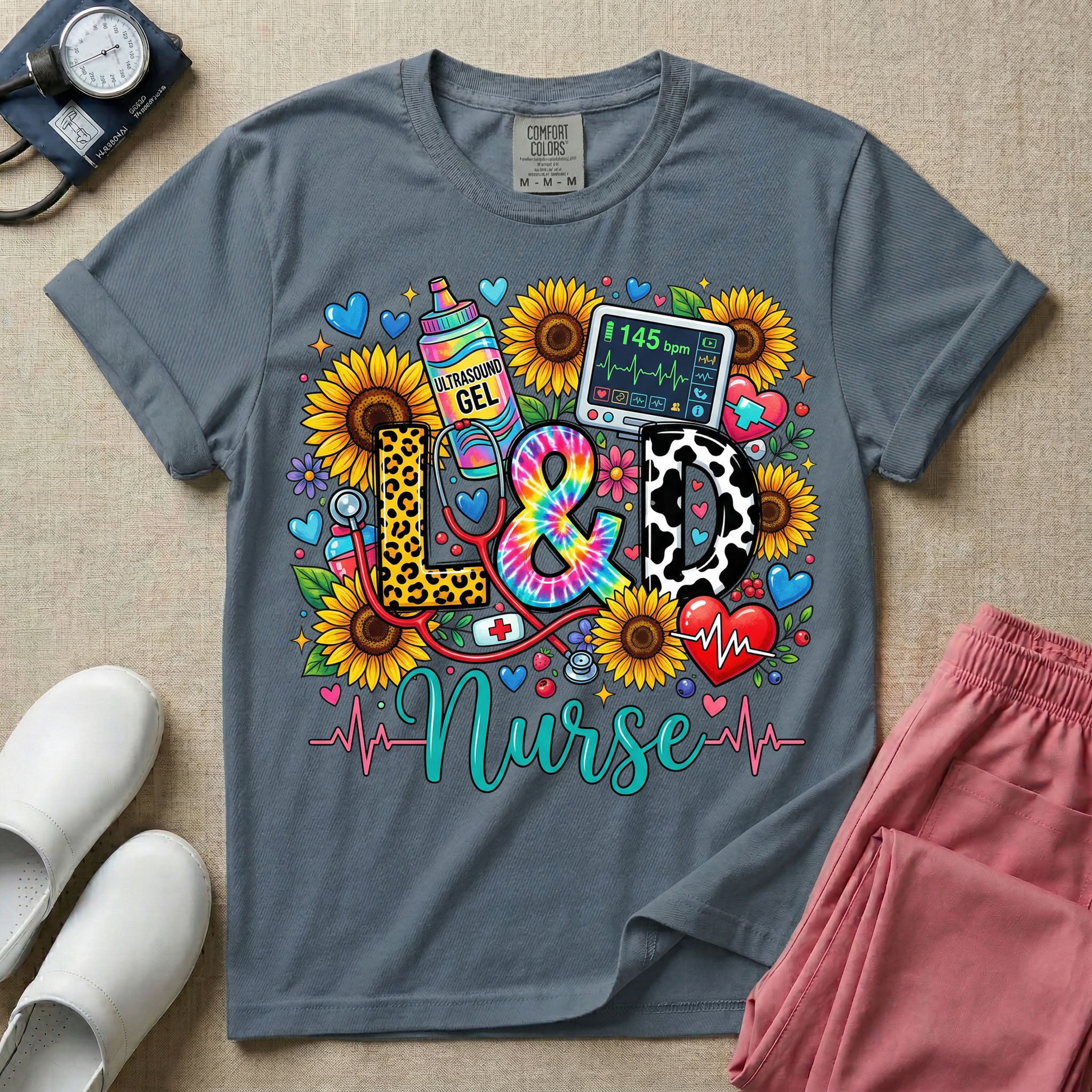 A colorful graphic t-shirt for a Labor and Delivery unit featuring the text "L&D" in leopard print, tie-dye, and cow print letters above the word "Nurse" in teal script, surrounded by detailed illustrations of sunflowers, a rainbow ultrasound gel bottle, a fetal monitor reading 145 bpm, a red stethoscope, and various hearts.