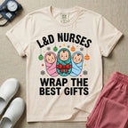A festive Christmas graphic t-shirt for a Labor and Delivery unit featuring the funny text 'L&D NURSES WRAP THE BEST GIFTS' surrounding an illustration of three cute babies swaddled in pink, green with a red bow, and blue blankets, accented with holiday ornaments, snowflakes, and stars.