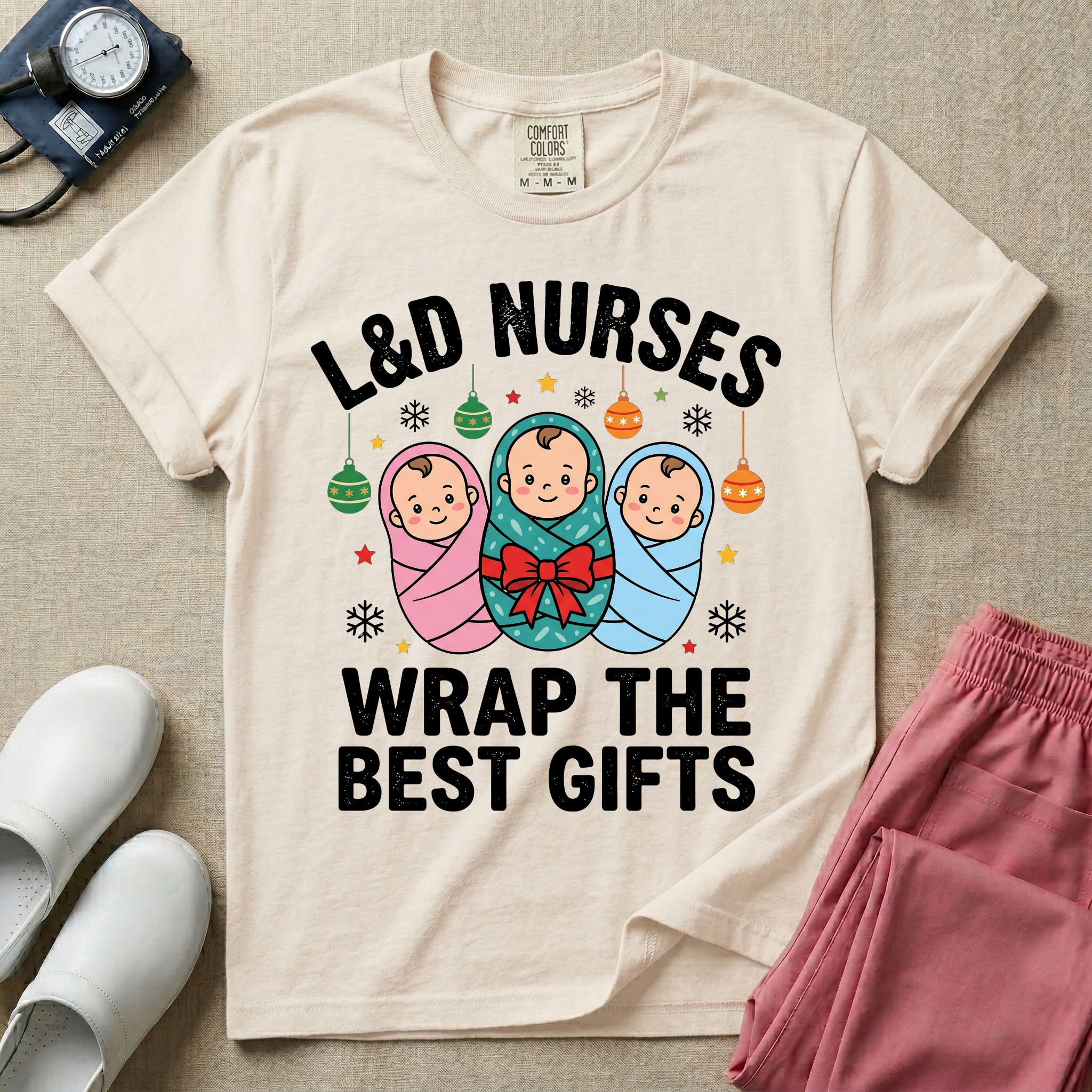 A festive Christmas graphic t-shirt for a Labor and Delivery unit featuring the funny text 'L&D NURSES WRAP THE BEST GIFTS' surrounding an illustration of three cute babies swaddled in pink, green with a red bow, and blue blankets, accented with holiday ornaments, snowflakes, and stars.