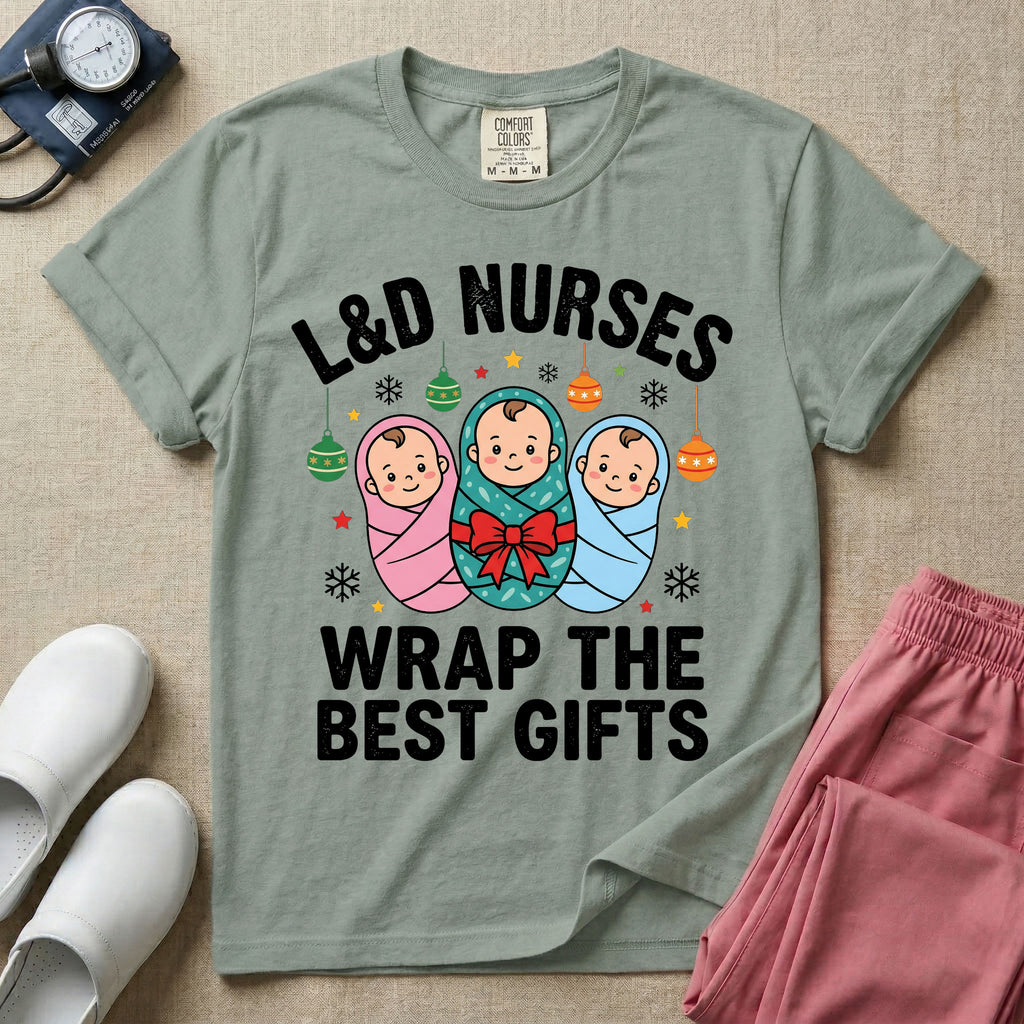 A festive Christmas graphic t-shirt for a Labor and Delivery unit featuring the funny text 'L&D NURSES WRAP THE BEST GIFTS' surrounding an illustration of three cute babies swaddled in pink, green with a red bow, and blue blankets, accented with holiday ornaments, snowflakes, and stars.