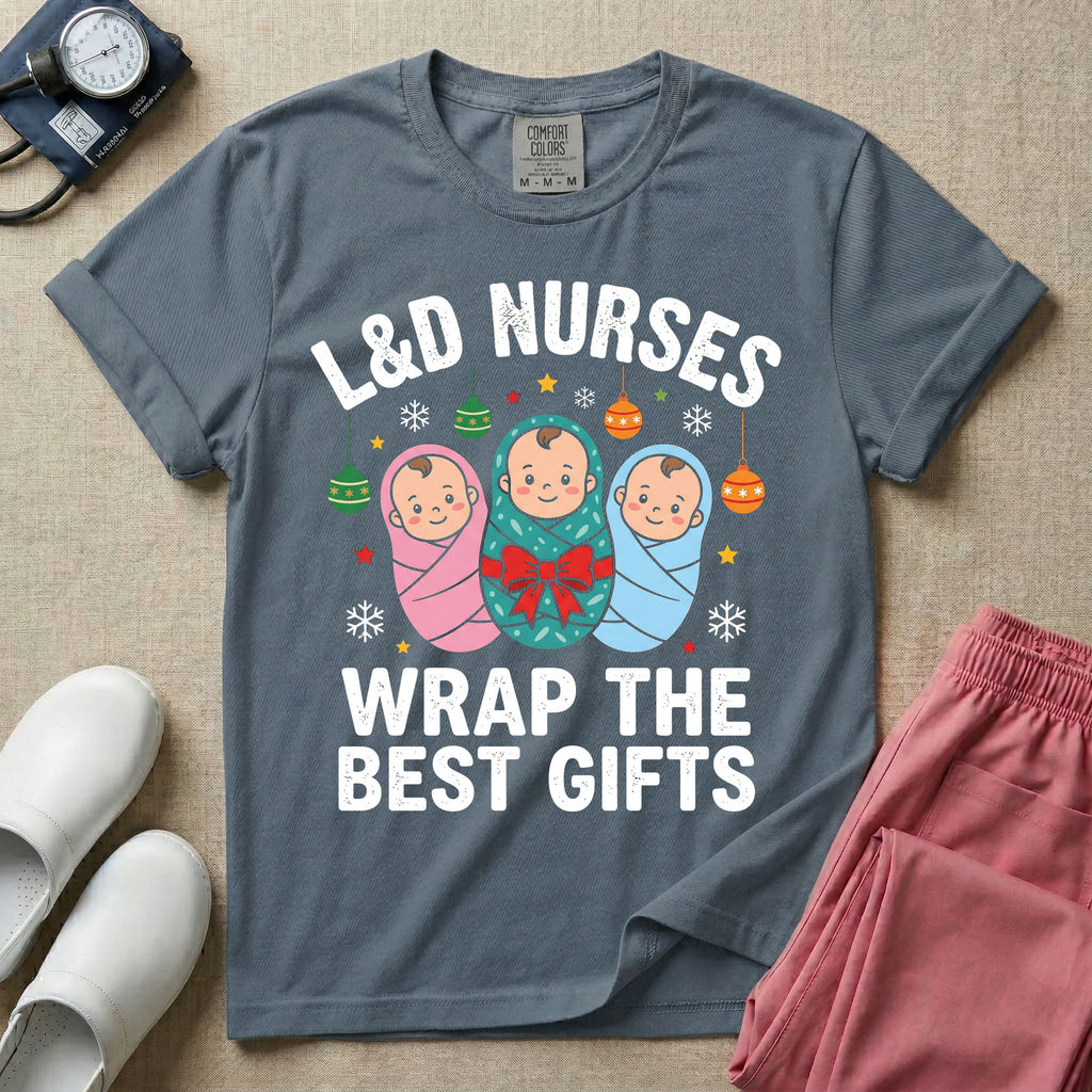 A festive Christmas graphic t-shirt for a Labor and Delivery unit featuring the funny text 'L&D NURSES WRAP THE BEST GIFTS' surrounding an illustration of three cute babies swaddled in pink, green with a red bow, and blue blankets, accented with holiday ornaments, snowflakes, and stars.