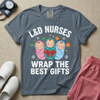 A festive Christmas graphic t-shirt for a Labor and Delivery unit featuring the funny text 'L&D NURSES WRAP THE BEST GIFTS' surrounding an illustration of three cute babies swaddled in pink, green with a red bow, and blue blankets, accented with holiday ornaments, snowflakes, and stars.