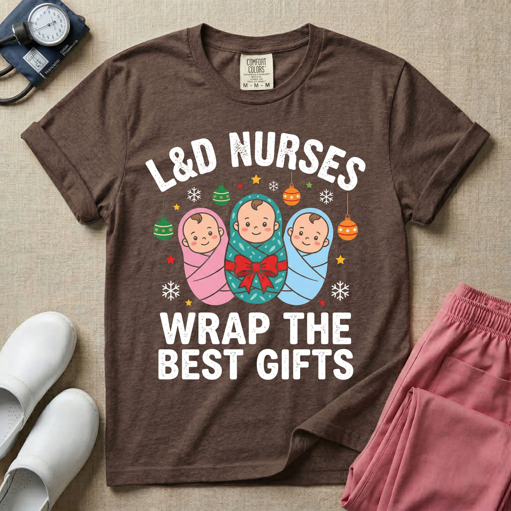 A festive Christmas graphic t-shirt for a Labor and Delivery unit featuring the funny text 'L&D NURSES WRAP THE BEST GIFTS' surrounding an illustration of three cute babies swaddled in pink, green with a red bow, and blue blankets, accented with holiday ornaments, snowflakes, and stars.