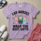 A festive Christmas graphic t-shirt for a Labor and Delivery unit featuring the funny text 'L&D NURSES WRAP THE BEST GIFTS' surrounding an illustration of three cute babies swaddled in pink, green with a red bow, and blue blankets, accented with holiday ornaments, snowflakes, and stars.