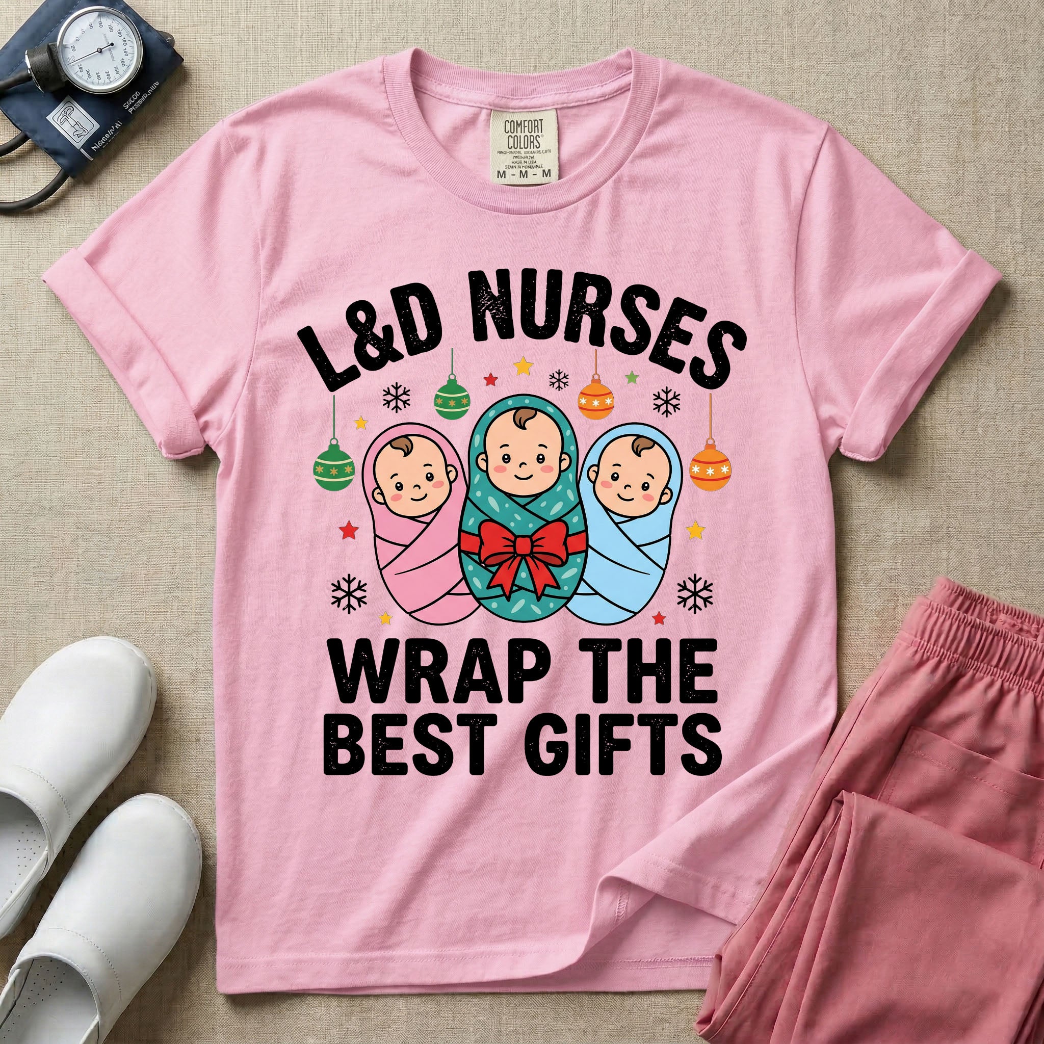 A festive Christmas graphic t-shirt for a Labor and Delivery unit featuring the funny text 'L&D NURSES WRAP THE BEST GIFTS' surrounding an illustration of three cute babies swaddled in pink, green with a red bow, and blue blankets, accented with holiday ornaments, snowflakes, and stars.