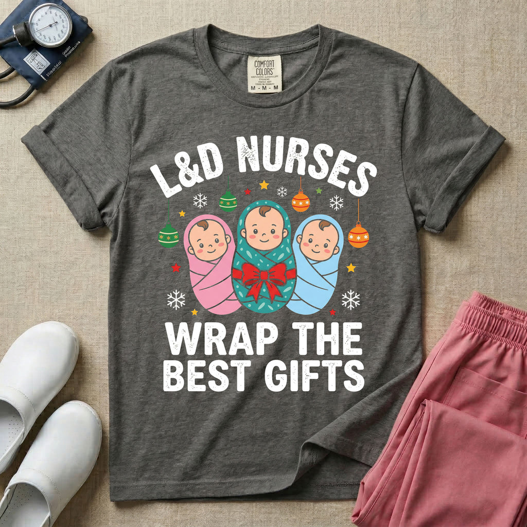 A festive Christmas graphic t-shirt for a Labor and Delivery unit featuring the funny text 'L&D NURSES WRAP THE BEST GIFTS' surrounding an illustration of three cute babies swaddled in pink, green with a red bow, and blue blankets, accented with holiday ornaments, snowflakes, and stars.