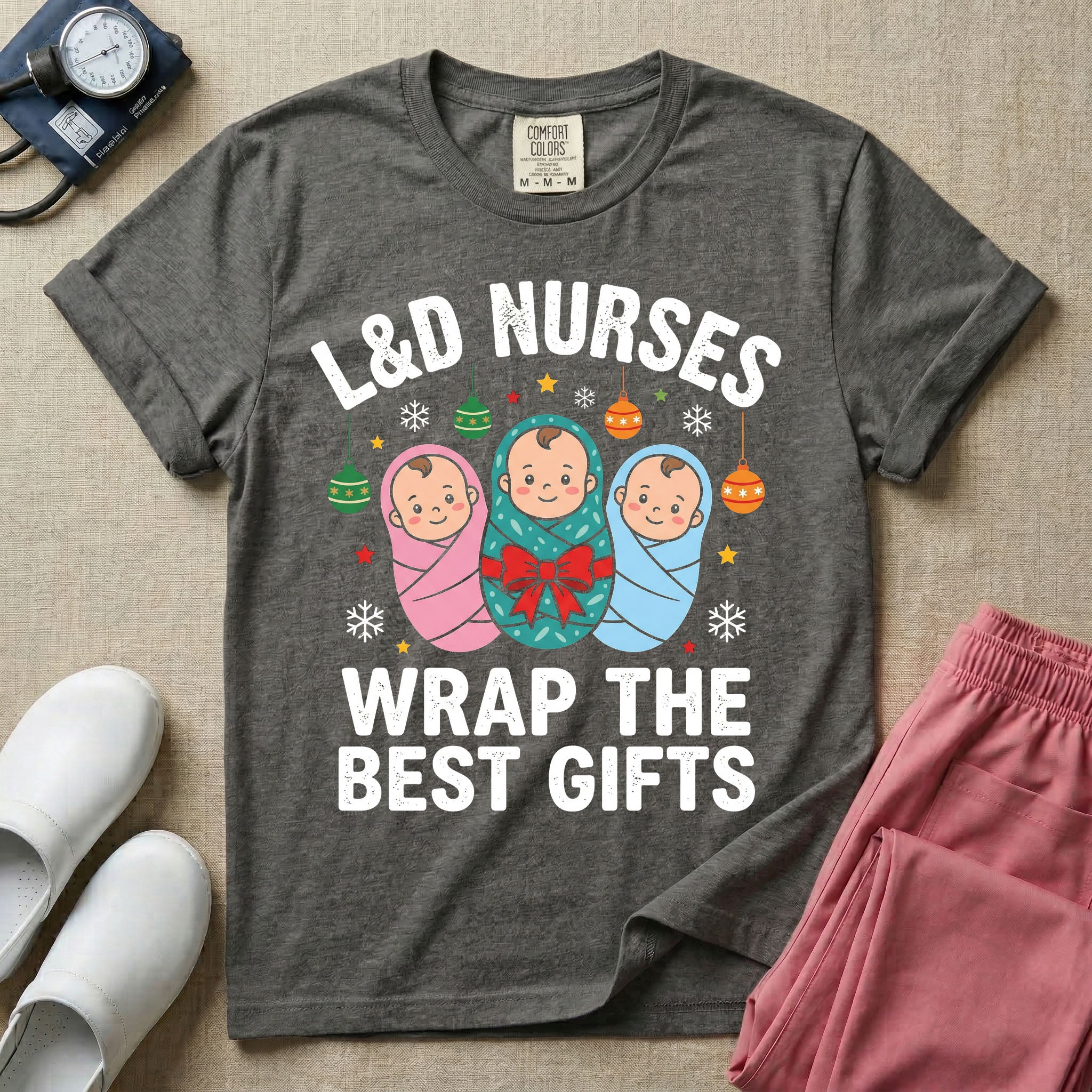 A festive Christmas graphic t-shirt for a Labor and Delivery unit featuring the funny text 'L&D NURSES WRAP THE BEST GIFTS' surrounding an illustration of three cute babies swaddled in pink, green with a red bow, and blue blankets, accented with holiday ornaments, snowflakes, and stars.