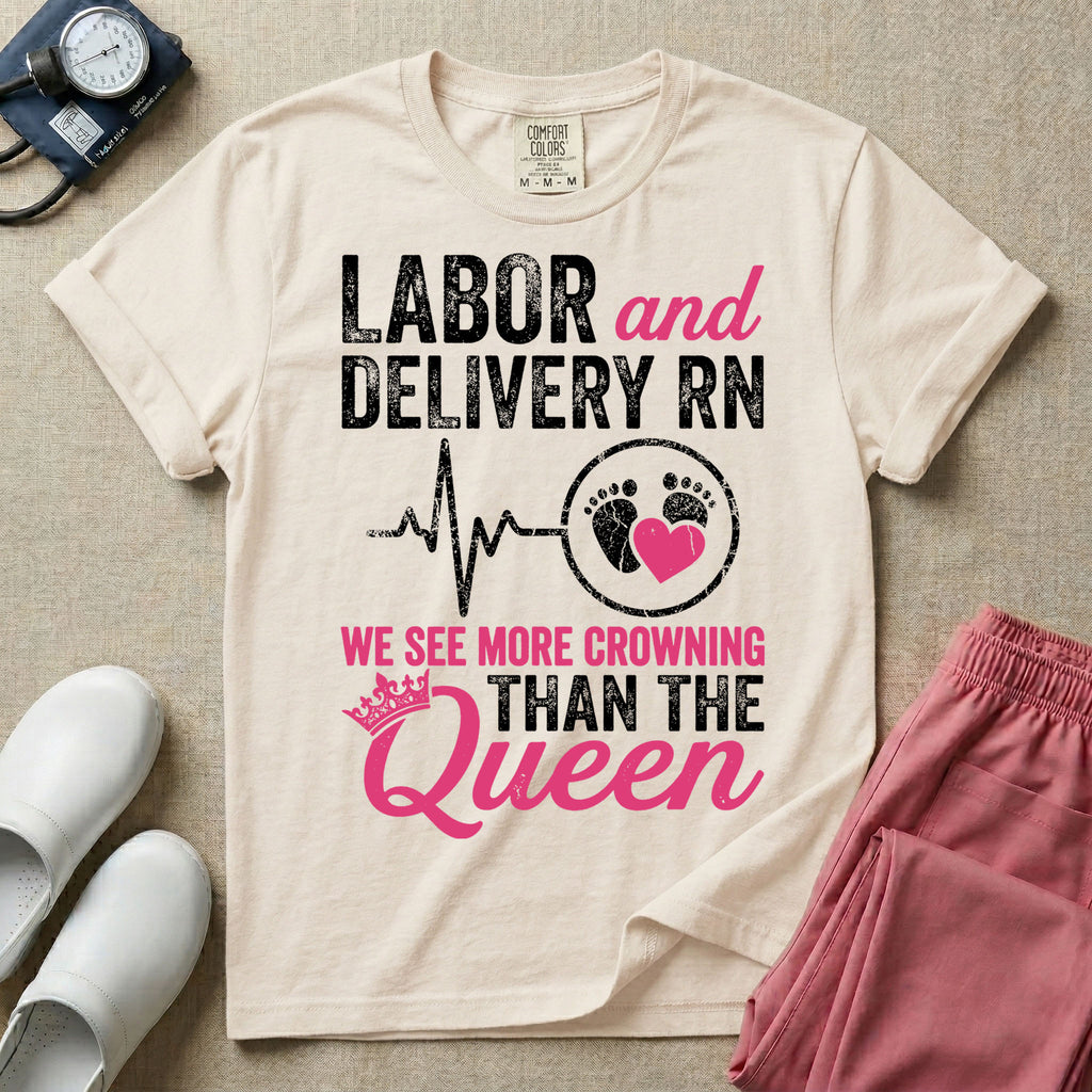 A funny graphic t-shirt for a Labor and Delivery unit featuring distressed white and pink typography that reads 'LABOR and DELIVERY RN' above a heartbeat line connecting to baby footprints and a pink heart, followed by the humorous phrase 'WE SEE MORE CROWNING THAN THE Queen' with a pink crown illustration.