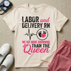 A funny graphic t-shirt for a Labor and Delivery unit featuring distressed white and pink typography that reads 'LABOR and DELIVERY RN' above a heartbeat line connecting to baby footprints and a pink heart, followed by the humorous phrase 'WE SEE MORE CROWNING THAN THE Queen' with a pink crown illustration.