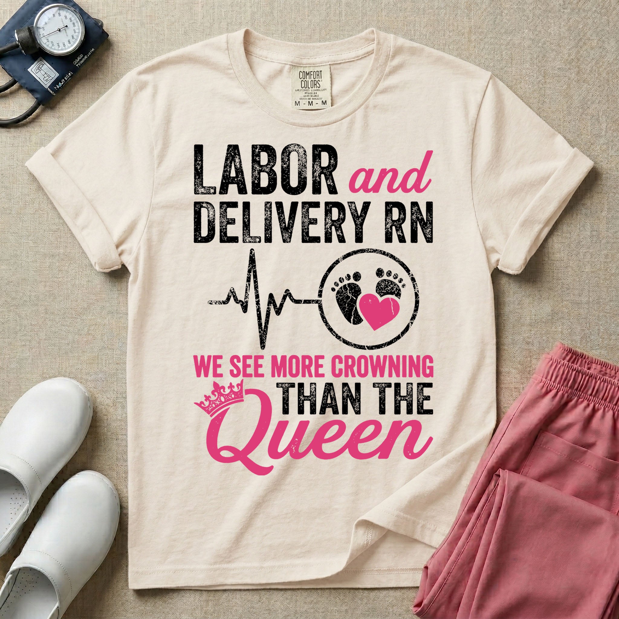 A funny graphic t-shirt for a Labor and Delivery unit featuring distressed white and pink typography that reads 'LABOR and DELIVERY RN' above a heartbeat line connecting to baby footprints and a pink heart, followed by the humorous phrase 'WE SEE MORE CROWNING THAN THE Queen' with a pink crown illustration.