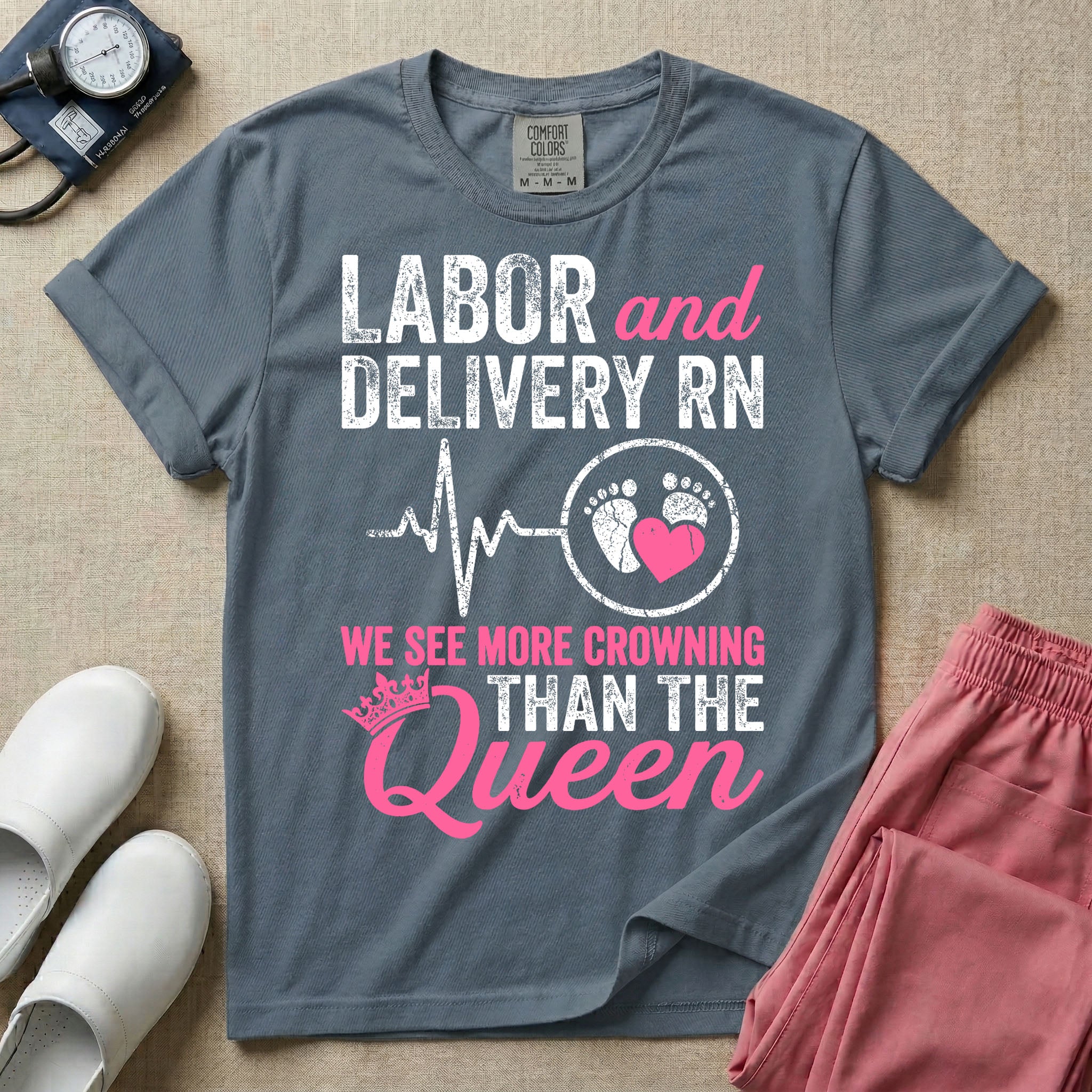 A funny graphic t-shirt for a Labor and Delivery unit featuring distressed white and pink typography that reads 'LABOR and DELIVERY RN' above a heartbeat line connecting to baby footprints and a pink heart, followed by the humorous phrase 'WE SEE MORE CROWNING THAN THE Queen' with a pink crown illustration.