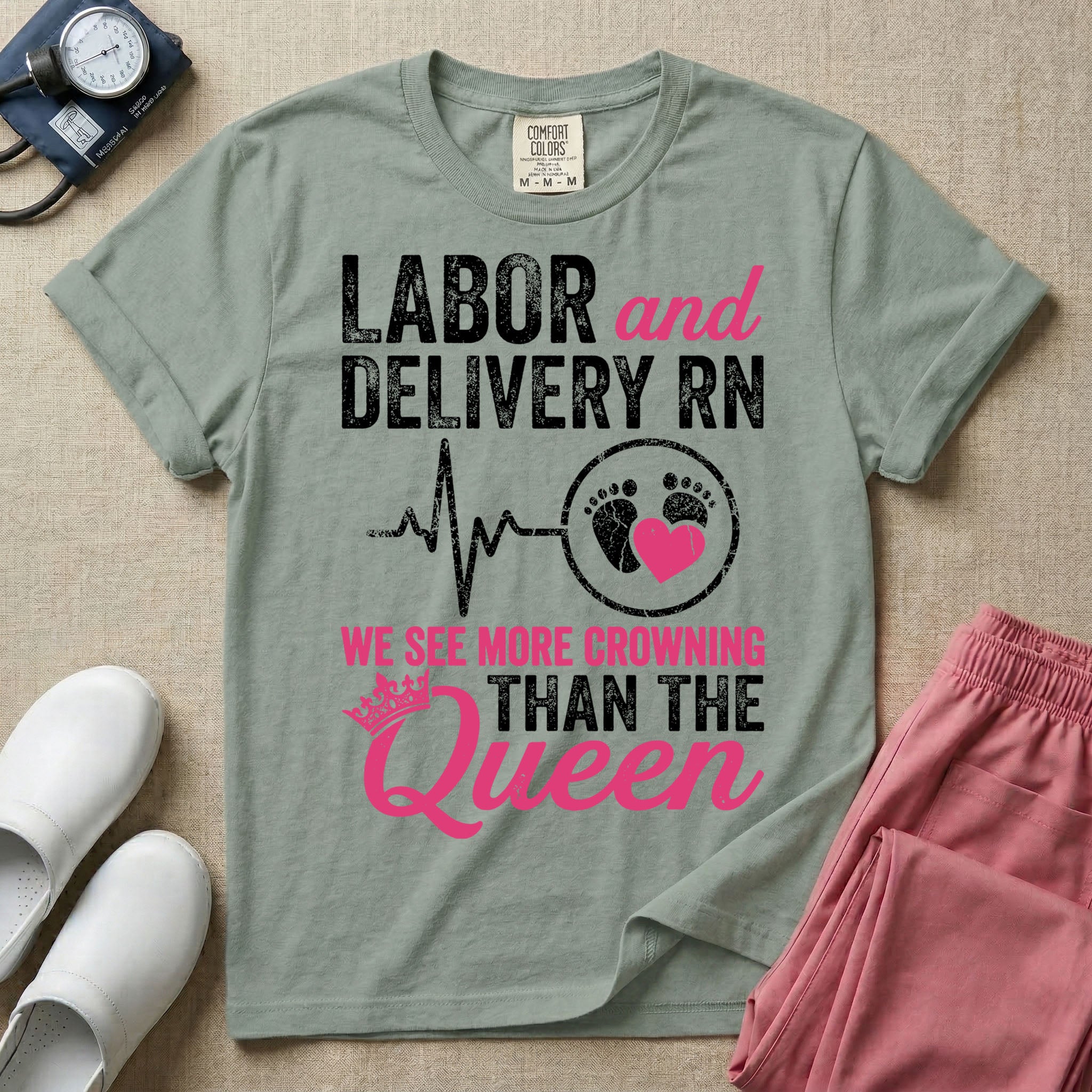 A funny graphic t-shirt for a Labor and Delivery unit featuring distressed white and pink typography that reads 'LABOR and DELIVERY RN' above a heartbeat line connecting to baby footprints and a pink heart, followed by the humorous phrase 'WE SEE MORE CROWNING THAN THE Queen' with a pink crown illustration.