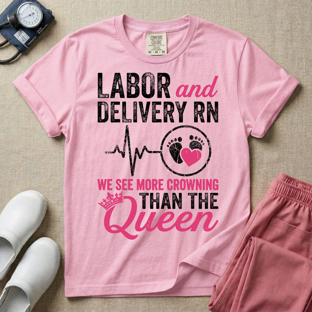 A funny graphic t-shirt for a Labor and Delivery unit featuring distressed white and pink typography that reads 'LABOR and DELIVERY RN' above a heartbeat line connecting to baby footprints and a pink heart, followed by the humorous phrase 'WE SEE MORE CROWNING THAN THE Queen' with a pink crown illustration.